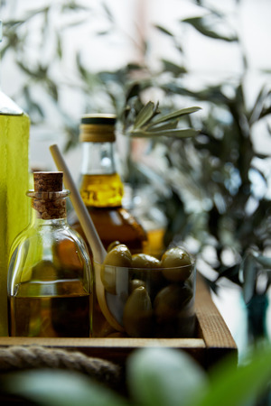 close up image of glass with spoon and green olives, bottles of aromatic olive oil with and branches on wooden trayの写真素材