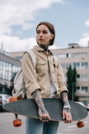 stylish tattooed woman looking away and holding longboard at streetの写真素材