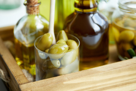 close up view of glass with spoon and green olives, jar, various bottles of aromatic olive oil on...
