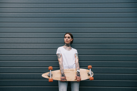 attractive young woman with tattoos holding skateboard against black wallの写真素材