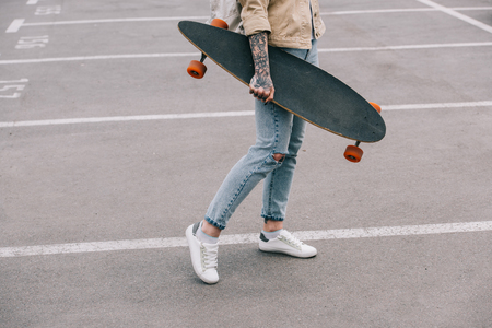 cropped image of stylish tattooed woman holding longboard at parking lotの写真素材