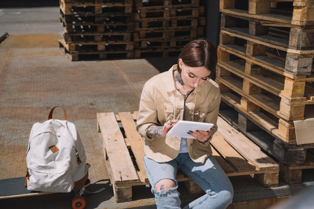 young tattooed woman using digital tablet near backpack and skateboard