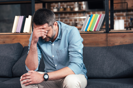depressed man with hand on forehead sitting on sofa in psychiatrist officeの写真素材