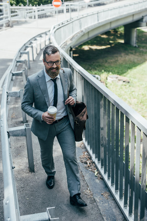 handsome businessman in formal wear with leather bag and coffee to go walking in cityの写真素材