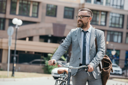 handsome businessman walking with bicycle on street in city and looking awayの写真素材
