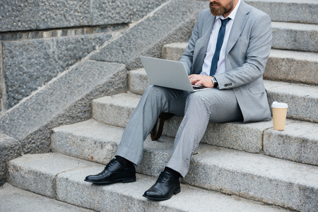 cropped view of businessman with disposable cup of coffee using laptop and sitting on stairsの写真素材