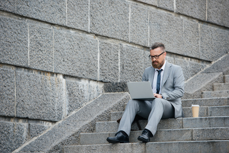 bearded businessman using laptop and sitting on stairs with coffee to goの写真素材