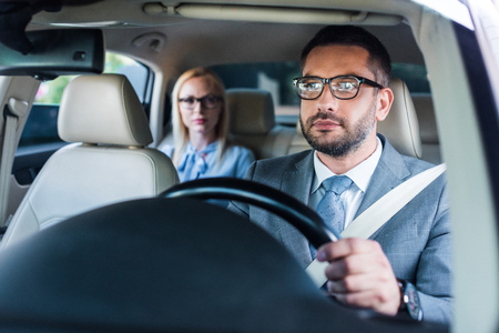 selective focus of businessman in eyeglasses driving car with colleague sitting behind on passengers seatの写真素材