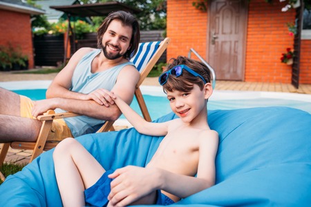 smiling father and son resting near swimming pool on backyard on summer dayの写真素材