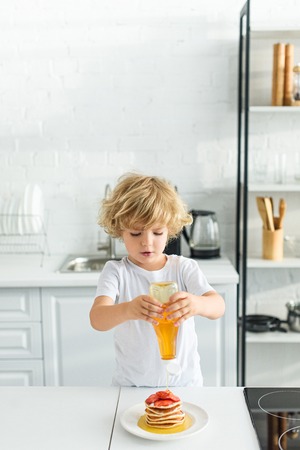 boy pouring syrup on pancakes with pieces of strawberry on plate at kitchenの写真素材