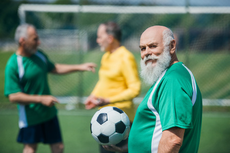 selective focus of old bearded man with football ball and friends behind on fieldの写真素材