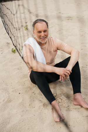 smiling elderly man with towel resting near net on sandy beach after playing volleyballの写真素材