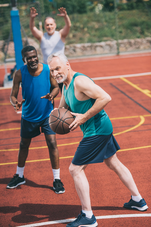 selective focus of old man with basketball ball in hands and multiracial friends behind on playgroundの写真素材