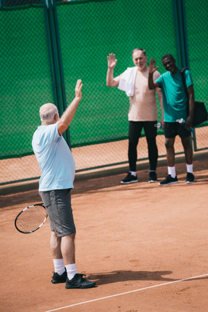 multiethnic old men greeting friend with tennis racquet on courtの写真素材
