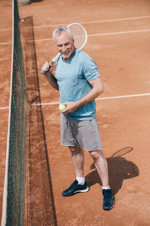 smiling elderly man in sportswear with tennis racquet and ball standing on courtの写真素材