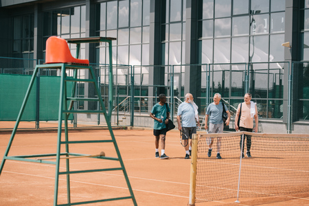 multiracial elderly friends with tennis equipment walking on courtの写真素材