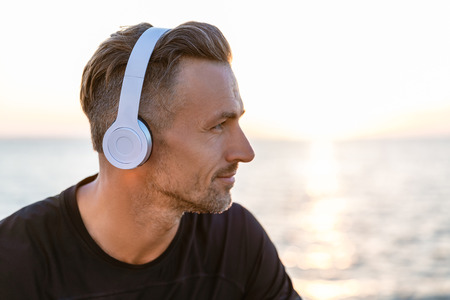 close-up portrait of handsome adult man in wireless headphones looking away on seashore in front of...