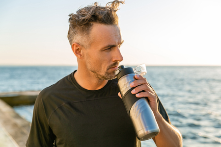 adult sportsman with grey hair holding fitness bottle on seashore in front of sunrise looking awayの写真素材