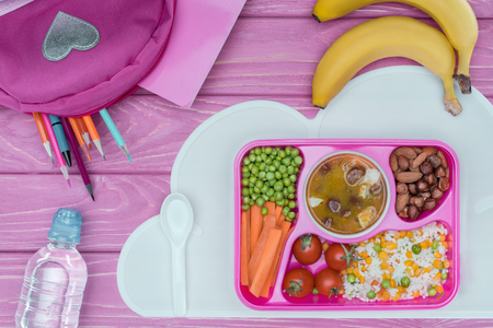 top view of tray with kids lunch for school, pink bag, pencils and bottle of water on pink tableの写真素材