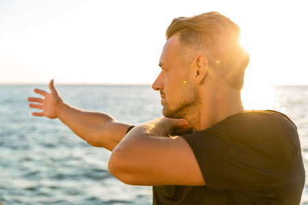 handsome adult sportsman stretching arm before training on seashore in front of sunriseの写真素材