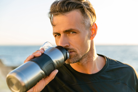 close-up portrait of handsome adult sportsman drinking water from fitness bottle on seashore in front of sunrise and looking at cameraの写真素材