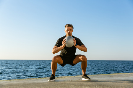 handsome adult sportsman doing squats with fit ball on seashoreの写真素材