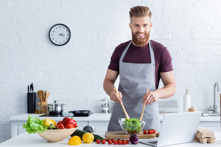 handsome bearded man in apron cooking vegetable salad and smiling at cameraの写真素材