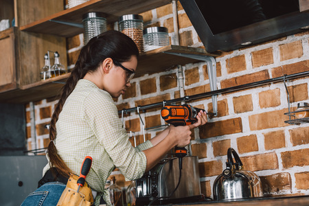 attractive young repairwoman in protective eyeglasses drilling wall at kitchenの写真素材