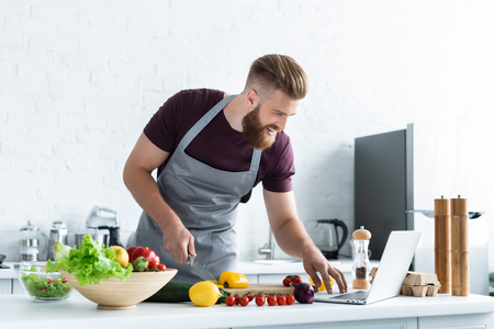 smiling bearded man in apron using laptop and cooking vegetable saladの写真素材