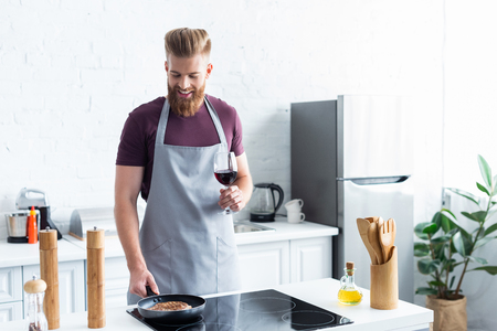 smiling young man in apron holding glass of red wine while cooking steak in frying panの写真素材