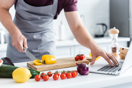 cropped shot of man in apron using laptop and cutting vegetablesの写真素材