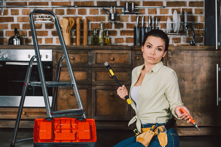 confused young repairwoman with hammer and pliers sitting on floor of kitchenの写真素材