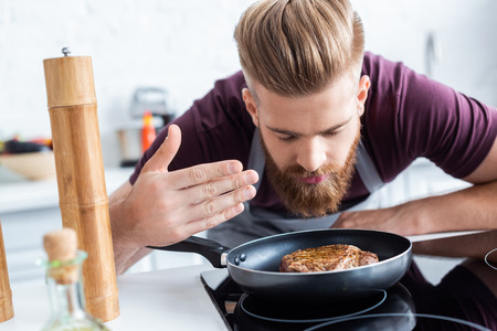 handsome bearded young man in apron cooking delicious steak on frying panの写真素材