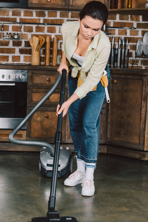 serious young woman with toolbelt using vacuum cleaner at kitchenの写真素材