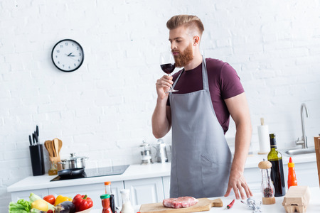 handsome bearded young man in apron tasting red wine while cooking in kitchen