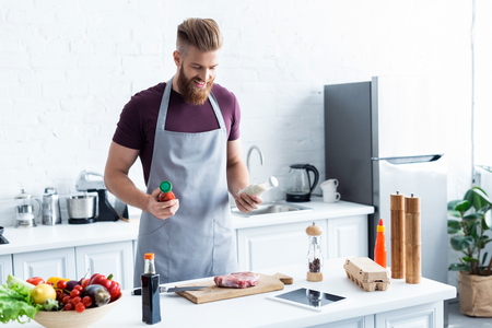 handsome smiling bearded man in apron cooking delicious steak in kitchenの写真素材
