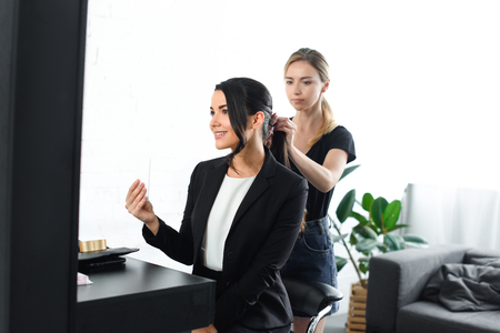 focused hairstylist doing hairstyle while smiling businesswoman in suit taking selfie in mirrorの写真素材