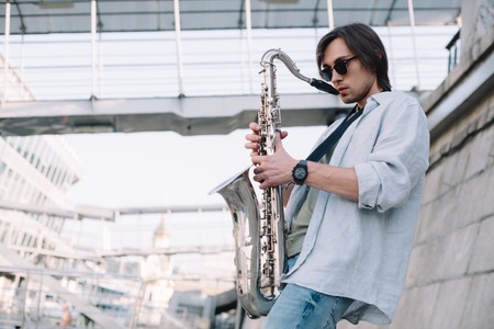 Young man in sunglasses adjusting saxophone on sunny city streetの写真素材