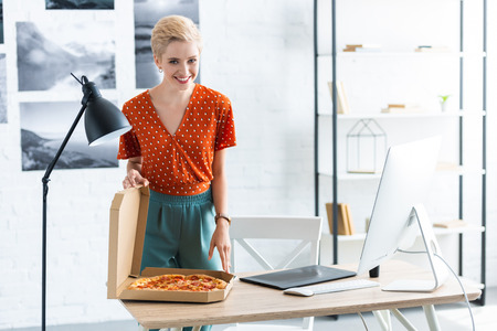 happy female freelancer opening pizza box at table with graphic tablet at home officeの写真素材