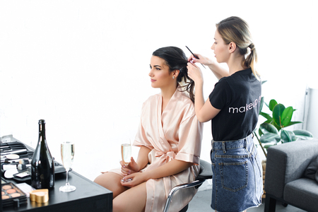 young hairstylist doing hairstyle for bride in silk bathrobe with glass of champagneの写真素材