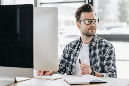 handsome young man in eyeglasses looking away while using desktop computer at workplaceの写真素材