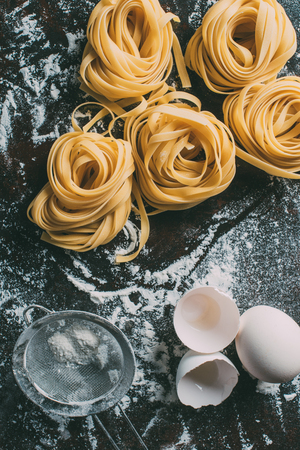 top view of raw tagliatelle pasta, sieve, egg and egg shell on table covered by flourの写真素材