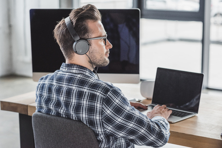 handsome young programmer in eyeglasses and headphones working with laptop and desktop computerの写真素材