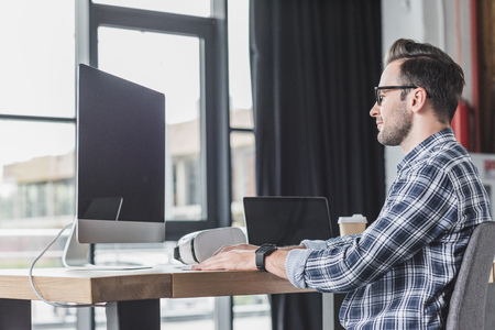 handsome smiling young programmer in eyeglasses working with desktop computer and laptopの写真素材