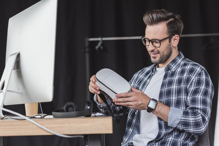 smiling young man in eyeglasses holding virtual reality headset at workplaceの写真素材