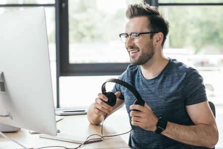 handsome smiling man in eyeglasses holding headphones and using desktop computerの写真素材