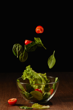 Tomatoes and salad leaves falling in bowl above wooden table surfaceの写真素材