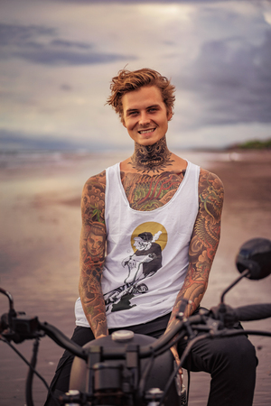 smiling handsome tattooed man sitting on motorcycle on ocean beachの写真素材