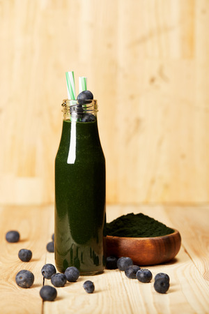 bowl with spiruluna powder, bottle of spirulina smoothie with blueberries and drinking straw on wooden tableの写真素材
