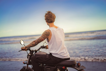back view of tattooed biker riding motorcycle on ocean beach